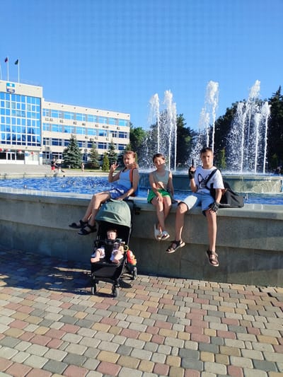 Three children and two infants are in the foreground of a sunny outdoor scene. A young girl with brown hair, wearing a light blue tank top and denim shorts, sits on the edge of a fountain and makes a "peace sign" with her fingers. Beside her in a stroller, two infants are visible; one is wearing a white onesie and the other a pink and white outfit. Next to the girl, a younger girl with brown hair wears a light green crop top and green shorts. To the right, a boy with short brown hair wears a white t-shirt, denim shorts, and black gloves, holding a black toy gun. He has a black bag on his shoulder. The children are sitting on the stone edge of a fountain with jets of water spraying into the air in the background. Behind the fountain is a large, modern, multi-story building with a blue-tinted glass facade and flags on top. The ground in front of the fountain is paved with colorful, interlocking rectangular stones. The sky is clear and blue. The scene appears to be in the city of Vinogradny, Russia, possibly in front of a government building or administrative center.