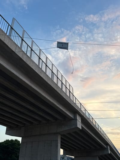 The image shows a concrete overpass with metal railings and wire mesh fencing. A white rectangular sign is hanging from a wire stretched across the sky. The sign has text in a dark color, with a lightning bolt symbol. Below the sign, a frayed wire hangs downwards. The sky is blue with scattered clouds, some of which are tinged with pink from the setting sun. Power lines are visible running parallel to the overpass. In the lower left corner, the top of a dark green tree is visible, and in the lower right, some buildings are partially obscured. The overpass is a significant piece of infrastructure, suggesting an urban or semi-urban environment. The sign's warning about touching wires and the danger to life indicates a potentially hazardous area, likely related to electrical infrastructure. The overall scene suggests a potentially unsafe but visually striking urban landscape at dusk.