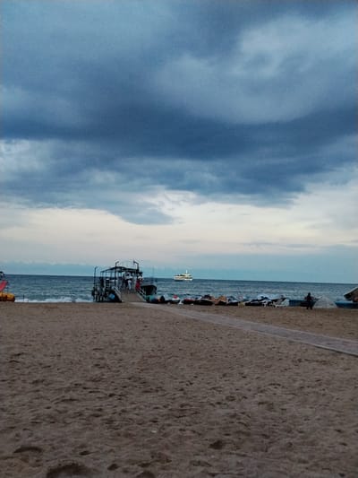 A tranquil beach scene unfolds under a dramatic, cloudy sky. The foreground is dominated by the textured sand of the beach, marked with the subtle imprints of footsteps. A pathway, made of light-colored material, leads towards the water.

In the middle ground, a pier juts out into the calm sea. Several people are gathered on and around the pier, with one boat docked alongside it. Beyond the pier, the sea stretches out to the horizon, meeting a sky filled with dense, dark blue clouds that gradually lighten to a soft gray and pale blue towards the horizon.

Further out at sea, a white passenger boat is visible, its form distinct against the dusky sky. Along the shoreline, various objects are scattered, including what appear to be small boats, some covered, and lounge chairs, suggesting a recreational area. A red and yellow structure, possibly part of a playground, is visible on the far left. The overall mood is serene, with the approaching twilight and overcast sky lending a slightly subdued, yet peaceful atmosphere to the setting. The location appears to be a coastal area, likely near a resort or a popular tourist spot.