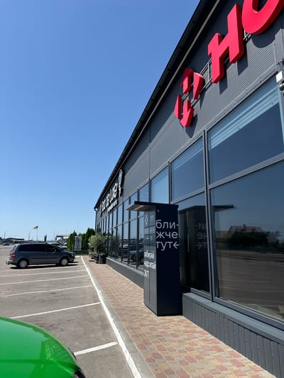 The image shows the exterior of a modern commercial building with large windows and a grey ribbed facade. Prominently displayed on the facade are large red three-dimensional letters that appear to spell "HO..." or a similar word. To the left of the building, a black parcel locker is visible, with Ukrainian text that translates to "Pick up, send". A parking lot with white lines is in the foreground, with a grey minivan parked in one of the spaces. A green vehicle's hood is partially visible in the lower left corner. In the distance, there is a Ukrainian flag on a flagpole and other buildings. The sky is clear and blue. This scene is located in the city of Sarny, Ukraine.