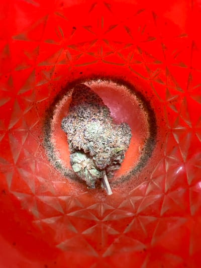 A single dried cannabis bud is centrally positioned within a bright red, textured container, viewed from a top-down perspective. The bud, approximately 3-4 cm in length, displays a dense structure with prominent green and purple coloration, heavily coated in visible trichomes, and includes a short stem. The container's interior surface features an embossed geometric pattern of interlocking triangles. A dark, granular residue, consistent with botanical material, forms a distinct circular accumulation on the container's floor, encircling the immediate area where the bud rests. No individuals, actions, or interactions are present. The specific location within the city of Hofit, Israel, is not discernible from the visual information provided in the image.