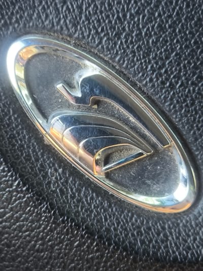 The image shows a close-up of a car's emblem, likely on the exterior or interior of a vehicle, set against a textured black background. The emblem is oval-shaped with a chrome finish that reflects light, suggesting it is made of polished metal. The emblem has a stylized design with curved lines and layered elements, giving it a sleek and modern appearance. The black background has a pebble-like texture, common on car dashboards or exterior trim. There are visible scratches and dust on the emblem and surrounding surface, indicating the car has been used. No people, specific activities, or emotional cues are depicted. The lighting suggests it is daytime, with strong reflections indicating a light source nearby. The location context provided is Abay, Kazakhstan, but no specific geographical or architectural cues in the image itself confirm this. No text is visible in the image.