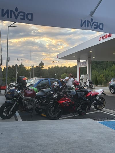 In the city of Unknown, Bulgaria, at a Lukoil gas station, several motorcycles are parked under a white canopy. The sky is filled with dramatic clouds at sunset, casting a warm glow. A man in a white t-shirt is taking a photo with his phone. The motorcycles include a red and black sportbike and a maroon and black naked bike. Two cars are also visible in the background. A sign with Cyrillic text and a logo indicating "Lukoil" is prominently displayed on the canopy. Another banner with a reversed logo that reads "DEVIN" is stretched across the top of the frame.