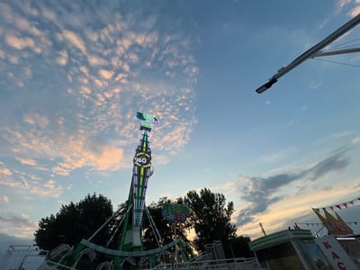 The image depicts an amusement park ride, a tall, green, and white structure with flashing lights, against a dusky sky with scattered clouds tinged with pink and orange hues. The ride appears to be a spinning thrill ride, with a circular sign displaying "360" prominently on its side. In the foreground, there are white railings and the lower portion of the ride's structure, also in green and white. Trees are visible behind the ride, silhouetted against the sky. A flag banner with colorful triangular flags is strung above a small building to the right of the ride. This scene is located at the amusement park in Golubitskaya, Russia. There are no people clearly visible in the image.
