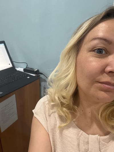 A woman with blonde hair and blue eyes is positioned in front of a laptop, which is on a wooden desk. A document rests next to the laptop. The background is a light blue wall. The location is not discernible as being in Beloretsk, Russia based on the image alone.