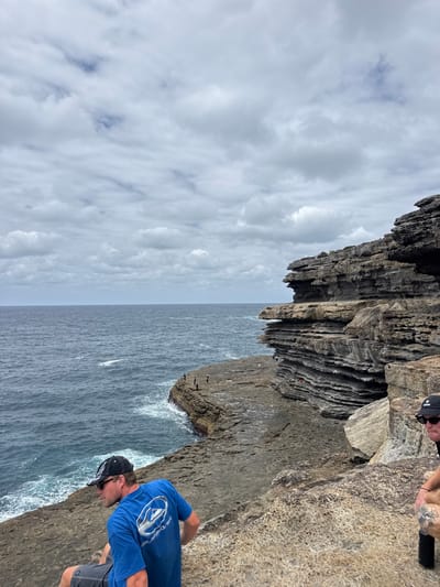 A rugged coastline dominates the scene, featuring layered rock formations meeting the ocean. The sea stretches out to a distant horizon, under a cloudy sky. A man in a blue shirt and cap stands on a cliff edge. Another person's hands are visible at the top of the frame. Below, tiny figures are present on a rock outcrop near the water's edge. Waves crash against the rocks.