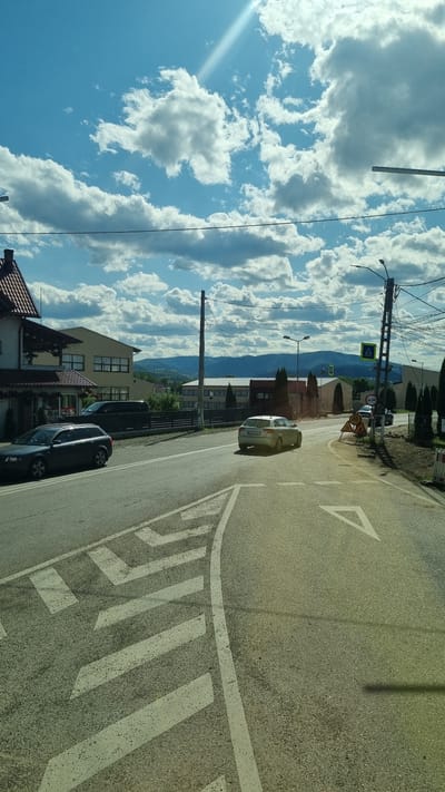 The image captures a bright, sunny day in Vicovu de Sus, Romania, with a partly cloudy sky. The foreground features a road with white directional markings. On the road, a dark sedan is parked on the left, and a silver hatchback is driving away from the viewer on the right. Beyond the road, there are several buildings, including a house with a red roof and a commercial building. In the background, rolling hills covered in trees are visible under the expansive sky. Traffic signals and streetlights are present along the road. Construction or road work appears to be ongoing on the right side of the road, indicated by barriers and signage.