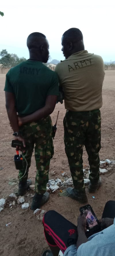 Two African soldiers in camouflage pants and "ARMY" labeled shirts stand facing each other outdoors. The soldier on the left wears a dark green shirt, while the other wears a tan shirt. One soldier holds a device with wires. A third person is partially visible at the bottom, looking at a phone screen. The background shows a dusty landscape.