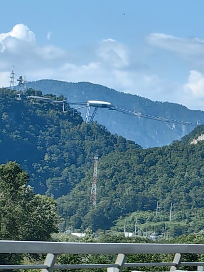 The image depicts a scenic mountain landscape with a complex cable car or gondola system traversing the slopes. The primary subjects are the natural environment and the man-made structure.
The scene is set on a clear, sunny day with a bright blue sky dotted with fluffy white clouds. The mountains are densely covered in lush green trees, suggesting a warm season, likely summer or late spring. In the foreground, a modern metal railing hints at a viewpoint or elevated walkway from which the photograph was taken.
The most prominent object is the cable car system, which appears to consist of multiple cabins suspended from cables stretched between towers on the mountain ridges. One station or terminal building, a sleek white structure with a curved roof, is visible on a higher peak. The system seems designed to transport people up and down the steep terrain, offering panoramic views of the surrounding natural beauty.
Other notable details include power lines and tall transmission towers visible on some of the peaks, indicating infrastructure development in the area. A tall, lattice-style tower, possibly a radio or cell tower, stands partway up one of the mountainsides. In the far distance, layers of mountains fade into a hazy blue, emphasizing the vastness of the landscape.
The overall mood is one of awe and tranquility, highlighting the juxtaposition of human engineering with the grandeur of nature. There is no visible text in the image. The location context provided is Kazachiy Brod, Russia.