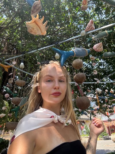 This is a selfie taken outdoors, possibly in a garden or courtyard, as indicated by the presence of lush green trees and some architectural elements in the background. The subject is a young woman with blonde hair, looking towards the camera. She is wearing a white top with a pattern of red cherries and a light-colored scarf around her neck.
The scene is decorated with a variety of objects hanging from what appears to be a metal frame structure, likely part of the outdoor decor. These objects include numerous seashells, coconuts, and some fish-shaped ornaments made of what looks like fabric or other materials, some wrapped in netting. The overall arrangement gives a whimsical, perhaps coastal, theme to the location.
The lighting suggests it is daytime, with bright sunlight casting shadows, hinting at a sunny day. The woman's expression is neutral, and her gaze is direct, typical of a selfie. Her left arm is visible, and she has a tattoo on her forearm. The background shows more of the decor and hints of buildings and greenery, consistent with a warm, possibly tropical, environment. The photo was likely taken in Tuy An Nam, Vietnam, based on the location context provided.