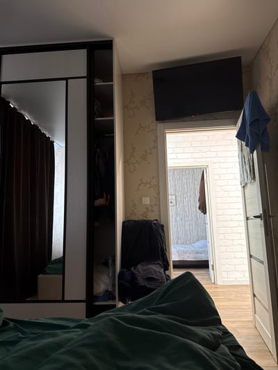 The image depicts an indoor scene, likely a bedroom. A large closet with mirrored doors and open shelves is visible on the left. The shelves contain various items, including clothing and storage boxes. To the right of the closet, a doorway leads into another room where a bed is visible. A television is mounted on the wall above this doorway. A blue towel hangs on the right side of the door frame. The foreground of the image is dominated by rumpled green bedding. The walls are decorated with wallpaper featuring a floral pattern. The flooring appears to be wood laminate. The lighting suggests it is daytime, with light entering from an unseen window or source, casting shadows. The scene is consistent with a residential interior.