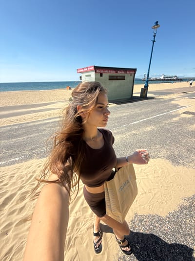 A young woman with long brown hair stands on a sandy beach in Bournemouth, United Kingdom, holding a Primark shopping bag. She is wearing a brown crop top and shorts, and black sandals. Behind her, a blue lamppost stands tall, and further in the distance, a pier extends into the ocean. The sky is clear and blue, and the sun is shining brightly. The scene appears to be a typical summer day at the seaside.