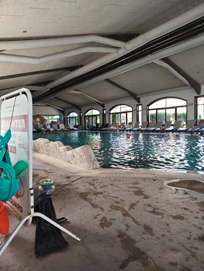 The image depicts the interior of a swimming pool facility. People are lounging on chairs along the poolside. In the foreground, there's a lifeguard stand with a lifeguard sign written in a Cyrillic alphabet (likely Bulgarian). Various swimming accessories such as goggles and flippers are visible near the lifeguard stand. The floor around the pool is wet. The architecture features a vaulted ceiling with visible pipes and beams. The setting is Krasnovo, Bulgaria.