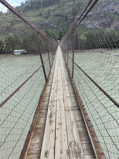 The image depicts a suspension bridge over a river. The bridge has a wooden plank walkway bordered by chain-link fencing supported by rusty metal poles and cables. The river appears murky and greenish, with gentle ripples. In the background, a steep, tree-covered mountainside rises, with a cloudy sky above. On the far bank of the river, partially visible through the fencing, is a light-colored van or bus. A person in the distance is walking on the bridge. The scene is likely outdoors, possibly in a rural or natural setting.