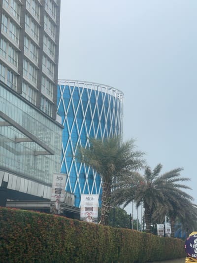 The image depicts a view of modern buildings and vegetation in Pakulonan Barat, Indonesia. On the left, a tall, rectangular building with a facade of many windows dominates the frame. Adjacent to it, to the right, is a distinctive cylindrical building with a blue glass exterior and a white diamond-shaped lattice pattern. In the foreground, a meticulously maintained hedge runs horizontally across the bottom of the image. Several palm trees are visible, with their fronds reaching upwards and obscuring parts of the buildings. Banners advertising "Atria Elegant Javanese" are placed in front of the hedge and near the buildings. The sky is overcast, indicating a cloudy day. The scene appears to be a street view, possibly from a vehicle, given the perspective and the presence of the hedge suggesting a roadside. There are no people clearly visible in the frame.