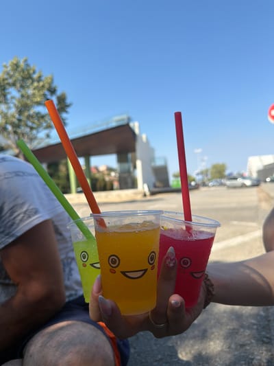 In Santa Susanna, Spain, a close-up shot captures three plastic cups with cartoon faces filled with brightly colored beverages and straws. The cup on the left contains a lime-green drink, the middle cup a vibrant orange drink, and the right cup a deep pink beverage. The cups are held by a person whose hand, with meticulously manicured nails, is visible in the foreground. The background is slightly blurred, showing a clear blue sky, trees, and a building structure, likely an outdoor recreational area or a part of the resort infrastructure. The scene appears to be outdoors on a sunny day, with some shadows visible on the ground, suggesting it's daytime.