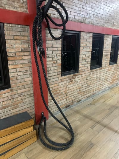 A black battle rope is coiled and lying on the floor of a gym in Kecamatan Kartasura, Indonesia. It is attached to a red wall with a metal loop. The rope is about 1.5 meters long and is made of thick, heavy-duty nylon. The gym floor is made of wood tiles. Three windows, two of them long and one shorter, are on the wall. One window has a metal frame. There are no people in the image. The location is inferred from the provided latitude and longitude.