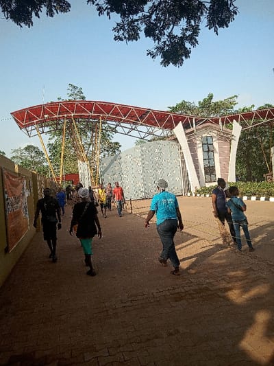 The image shows a group of people walking through an entrance in the city of Otta, Nigeria. The entrance is adorned with a red and grey steel structure with a canopy above. The people are walking on a paved pathway that leads to a larger paved area beyond the entrance. The sky is clear and bright. There are trees and foliage surrounding the entrance. The group of people appear to be entering or leaving a venue or building. One woman wearing a blue t-shirt and black pants is leading the way. Some people are walking in pairs, while others walk alone.  The ground is a mix of brick and dirt. There are yellow caution lines painted on the ground.