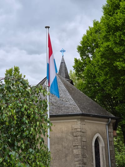 The image shows a section of the building of the European Museum Schengen in Luxembourg. The Luxembourg flag is displayed on a flagpole in front of the building. The building has a grey stone exterior with a gothic arched window. The roof is slate tile, and a blue cross is atop a steeple. The building is surrounded by greenery. The sky is cloudy.
