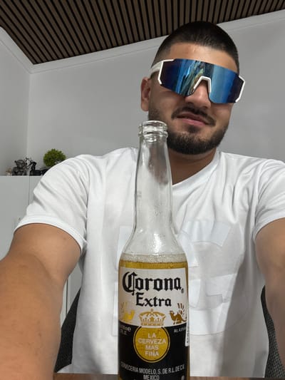 In the city of Rosenovo, Bulgaria, a young man with a short haircut, beard, and mustache is holding a bottle of Corona Extra beer. He is wearing a white t-shirt and large, futuristic white sunglasses with blue mirrored lenses. The bottle is mostly in focus and positioned in the foreground, obscuring part of the man's face and body. In the background, a white wall is visible, and above it, a ceiling with dark horizontal slats. To the left of the man, a shelf holds a small decorative monkey figurine and a green shrub. The lighting suggests an indoor environment, possibly an office or a home setting.