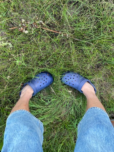 A first-person perspective shot from a standing position shows a person wearing blue denim shorts and dark blue Crocs, standing on a patch of green grass with some dry twigs and leaves scattered on the ground. The location is identified as Oktyabr'skii, Russia.