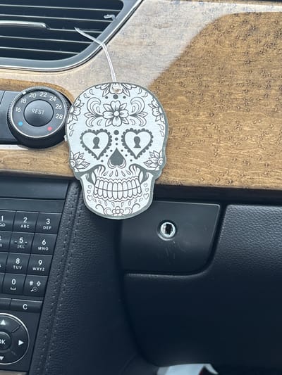 The image shows a close-up view of a car's interior dashboard. In the center, a skull-shaped air freshener with decorative floral patterns is hanging from an air vent. To the left of the air freshener is a circular dial with temperature settings ranging from 16 to 28, labeled "REST" in the center. Below the air freshener and dial is a section of the car's center console, featuring a black, textured surface and a keypad with numbers and symbols. The background is dominated by the wood-grain texture of the dashboard. The scene is well-lit, likely by daylight. There are no people visible in the image. This scene is not in Гацукоўскі сельскі Савет, Belarus, as it depicts a car interior.