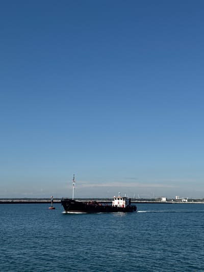 A long black ship travels in the blue Adriatic Sea off the coast of Ravenna, Italy. The vessel proceeds parallel to the coastline, generating a wake in the water. A blue sky dominates the landscape above the scene.
