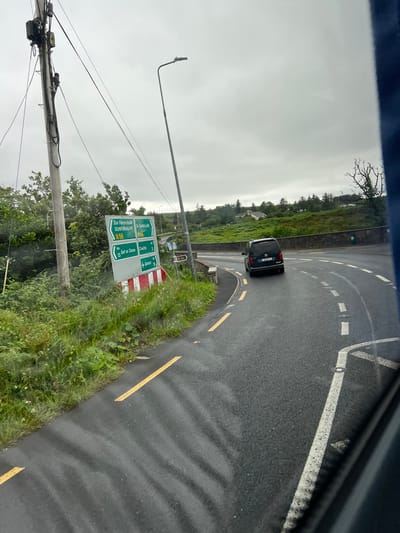 The image depicts a scenic view of a road in Gweedore, Ireland. A black van is driving on a wet asphalt road that curves to the left, bordered by lush green grass and trees on one side. The sky is overcast, suggesting a cloudy or rainy day. A signpost provides directional information to various locations, including Dunfanaghy, Gort an Choice, Croichli, and Derfort, with distances indicated in kilometers. The sign is primarily green with white lettering and features red and white stripes at the bottom. Electrical wires and a lamppost are visible to the left of the road, indicating a somewhat developed area within the natural landscape. The overall scene conveys a sense of travel and the picturesque, albeit gloomy, Irish countryside. The license plate on the van is partially visible but unreadable.