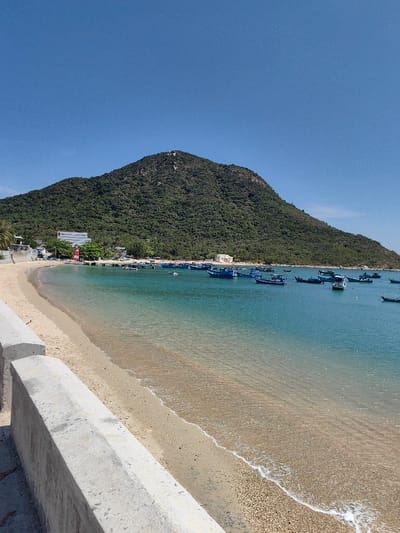 A sunny coastal view showcases a sandy beach meeting clear turquoise water, filled with numerous blue boats. A large, lush green mountain rises in the background, contrasting with the bright blue sky. A grey concrete barrier runs along the edge of the beach in the foreground. Buildings and a red and white lighthouse are visible near the shore at the base of the mountain.