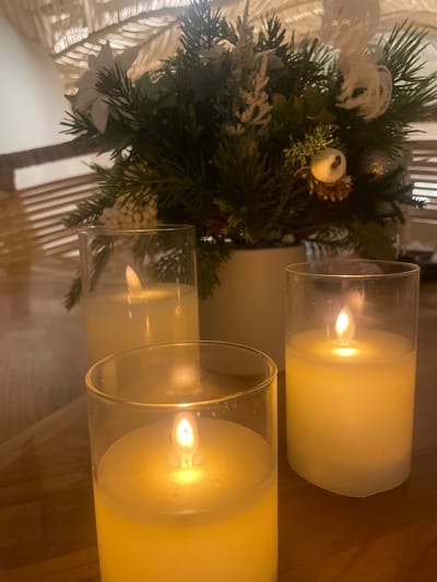 A dimly lit photograph captures three LED candles with flickering faux flames, each encased in a clear glass cylinder. The candles are arranged on a wooden surface, likely a table, with a festive floral arrangement in the background. The arrangement features pine branches, a white poinsettia, and small white berries, suggesting a holiday or winter theme. The overall ambiance is warm and inviting. The image does not contain any discernible elements to indicate its location in Gimpo-si, South Korea.