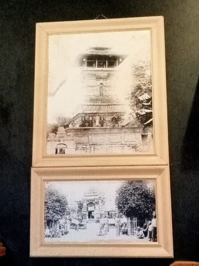 The image is of two black and white framed photographs hanging on a dark wall. The top photograph shows a large stone structure in the center of the image with smaller structures around the base. The structure appears to be a pagoda or temple. The top photograph appears to be older and is faded. The bottom photograph shows a newer image of the same building but from a wider perspective. A courtyard is in front of the structure and there are several people walking around. The coordinates provided place the location of the images in Kudus, Indonesia, at a historic site or religious place. The images appear to be from a collection of historical photographs of Kudus.