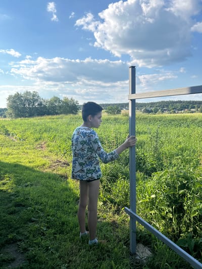 A young boy, viewed from the side, stands in a grassy field in Vyshevychi, Ukraine. He is wearing a long-sleeved shirt with a dinosaur pattern and dark shorts. His right hand rests on a vertical metal pole, part of a horizontal metal structure. The field is lush and green, with a path visible in the background. Trees and a distant village are visible on the horizon under a bright blue sky with scattered white clouds.