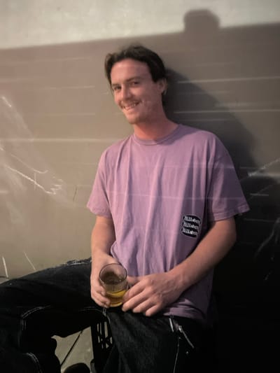 A male individual is depicted in a seated position against a light-colored wall, located in Thirroul, Australia. The man, with short dark hair and faint facial hair, is smiling directly at the camera. He is wearing a light purple, short-sleeved t-shirt featuring subtle horizontal striations and a black patch on the left chest with three stacked "Billabong" logos. His lower attire consists of dark jeans, exhibiting prominent seams and a light-colored zipper or chain detail on the right thigh. In his right hand, he holds a clear, ribbed glass containing an amber-colored liquid. His left hand rests near the base of the glass. The background consists of a textured wall, with a section on the left covered by a translucent, wrinkled plastic sheet. A dark, distinct shadow, consistent with a human head, is cast on the wall above the man's right shoulder. A dark object, possibly a plastic crate, is partially visible beneath him, supporting his seated position.