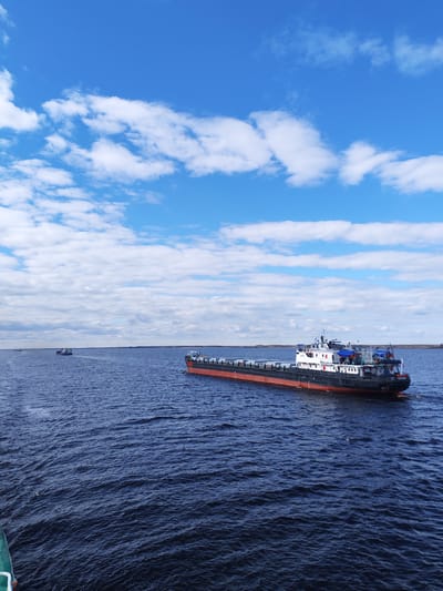The image displays a vast expanse of dark blue water, likely a large river or reservoir, under a bright, clear blue sky adorned with scattered white cumulus clouds. The setting is Maksakovo, Russia.

In the mid-ground, a long cargo vessel, possibly a self-propelled barge or a push-tug and barge, moves from left to right across the frame. The vessel features a red lower hull, black sides, and a white superstructure towards the stern, which is partially covered by blue tarpaulins or canvas. On the stern, the name "КОРСАР" (KORSAR) is clearly visible in white letters. The vessel creates a slight wake as it glides through the water.

Further in the distance on the left, a smaller vessel, appearing to be a tugboat, is also navigating the waterway. An even more distant, faint silhouette of another watercraft can be seen on the far-left horizon. The distant shoreline on both sides of the water is low and appears to be forested.

The photograph is taken from the perspective of another boat, with a partial view of a green hull or railing visible in the bottom-left corner. The time of day is clearly daytime, characterized by bright sunlight and fair weather. The scene conveys a sense of industrious activity on a wide, calm waterway.
