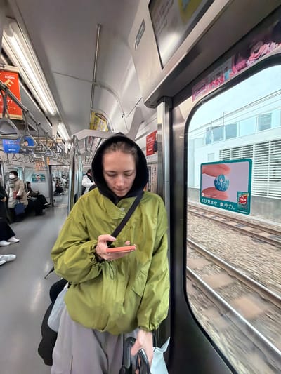 The image captures a candid moment inside what appears to be a Japanese commuter train, viewed from a slightly low angle, looking towards the front of the car. The original image is rotated 90 degrees clockwise, but I will describe it as if it were correctly oriented.
In the foreground, a person is seated or standing, looking downwards or with their eyes closed, giving an impression of rest or contemplation. They are wearing a hooded, light olive-green jacket over a dark-colored hoodie. A black strap, likely from a bag, crosses their chest. In their hands, they hold a light orange or pink smartphone, which has a small, light-colored charm (possibly a heart shape) attached to it. Their hair is dark brown.
Through the window to the right of the person, a blurred view of train tracks and an adjacent building with multiple windows can be seen, indicating the train is in motion. A sticker is affixed to the window, featuring an image of hands holding a small, stylized blue globe. Below the image, Japanese text reads "より覚ませ、集中力。" which translates to "Wake up more, focus." or "Awaken your concentration." There is also a small cherry blossom icon and other smaller Japanese characters, possibly product branding.
The interior of the train car is well-lit, likely by fluorescent lights, and features light-colored walls and ceiling. Overhead handrails extend along the length of the car, and several hanging straps are visible for standing passengers. Further down the train car, other passengers are visible, some of whom are wearing face masks. The overall ambiance suggests a typical daytime journey on public transportation. On some of the internal advertisements, Japanese text including the brand name "KIRIN" can be partially seen.