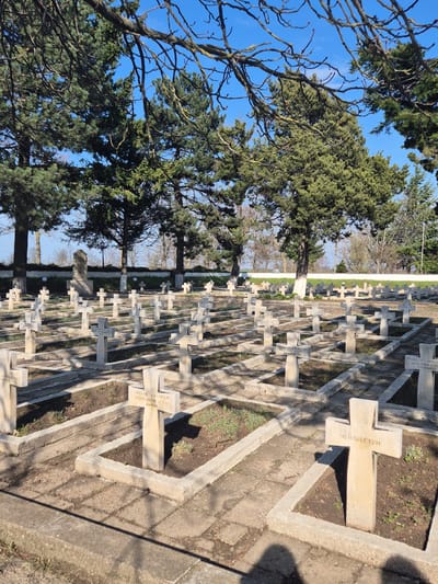 The image shows a cemetery with rows of stone crosses under a blue sky and trees in Shumen, Bulgaria. The crosses bear inscriptions in Cyrillic script, including the names of the deceased. The setting suggests a location of remembrance and respect for the deceased. The crosses are set in concrete enclosures with patches of dirt and greenery. The overall impression is one of order and reverence.
