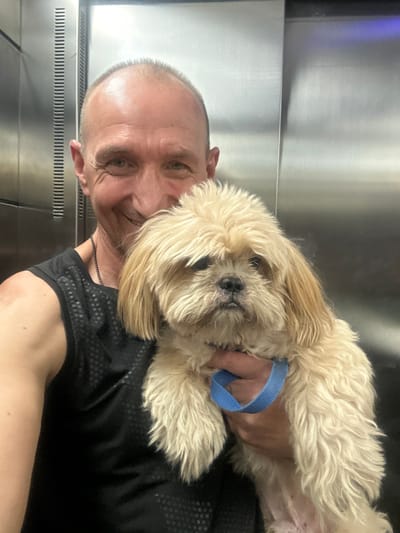 The image shows a close-up, selfie-style shot of a bald, light-skinned man in his 40s or 50s, holding a small, light-colored, fluffy dog. The man is wearing a black sleeveless athletic shirt and a thin chain necklace. He is smiling directly at the camera. The dog, which appears to be a Shih Tzu or similar breed, has its head resting on the man's arm. It has light brown and blonde fur and is wearing a blue collar or leash attachment. They are standing in what appears to be an elevator, with the walls of the elevator being metallic and reflective. The background shows vertical lines typical of elevator interior panels. There is no indication of the city of Arad, Israel in the image.