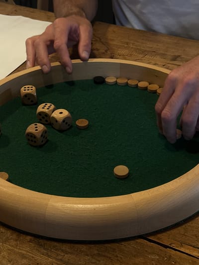 A person is playing a game on a circular wooden board with a green felt surface. The game involves wooden discs, some light and one dark, arranged around the edge of the board and scattered in the center, along with several dice. The person's hands are visible as they manipulate the discs. The board rests on a wooden surface, and a piece of white paper is visible on the left side of the image. The location is not evident from the image, and therefore cannot be associated with La Couarde-sur-Mer, France.