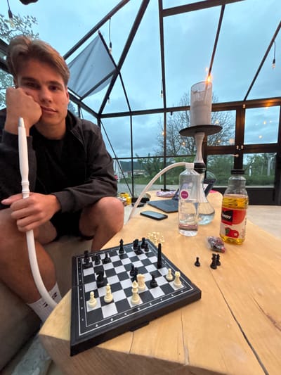 A young man is seated in a room with a glass roof and walls, likely a greenhouse or sunroom. He is holding the hose of a hookah in his hand. On a wooden table next to him is a chessboard with pieces set up, a laptop computer, two bottles of liquid, and other small items. The location is likely in Vlado Trichkov, Bulgaria. The hookah is in use, as evidenced by the burning substance at the top. The chess game appears to be in progress. The man is looking directly at the camera.