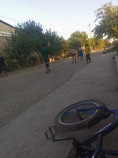 This media file depicts a street scene in Tahmach, Uzbekistan, likely during the late afternoon or early evening. The sky is clear and bright, suggesting fair weather. The foreground is dominated by a bicycle lying on its side on a gravelly, asphalt road. The bicycle's wheel has spokes of various colors. Several young people, seemingly boys, are scattered across the road and the side of the path. One boy in a green t-shirt and shorts is walking away from the camera. Other boys are standing and conversing. The background shows a residential area with houses and lush green trees lining the street. Power lines are visible overhead, and a utility pole stands in the distance. The overall atmosphere appears casual and relaxed, indicative of a typical neighborhood scene where children are playing or socializing outdoors. There is some text visible on the frame of the bicycle, which appears to read "BONVOY".
