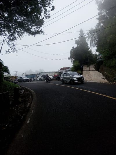 The image captures a foggy daytime scene on an asphalt road in Phoxay, Laos. The road, marked with a solid yellow line on its right side, curves to the left and ascends. A silver multi-purpose vehicle (MPV) is prominently positioned on the right side of the road, moving towards the viewer. Further back and to the left of the MPV, a dark-colored motorcycle carries two riders, also moving in the same general direction. Various other vehicles, including a white sedan and a pickup truck, are visible in the mid-ground, some appearing parked on the left side of the road next to a structure with a blue awning. Overhead power lines span across the upper portion of the frame against a hazy, light gray sky, indicative of heavy fog or mist. On the immediate right, a paved driveway leads uphill to a dark metal gate, where a single individual in a dark shirt stands. This area is flanked by a vegetated retaining wall and dense green foliage and trees extending up the steep embankment, which disappears into the fog. The left side of the road features a dark, overgrown shoulder.
