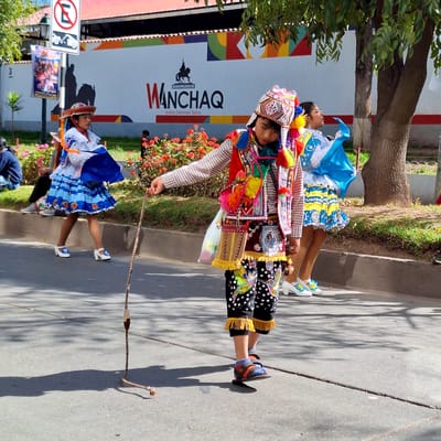 The image depicts a street scene in Wanchaq, Peru, likely during a parade or festival, as indicated by the traditional colorful attire of the individuals. In the foreground, a young person in a detailed costume, featuring a patterned hat with tassels, a vest adorned with embroidery and pom-poms, and patterned pants with fringed hems, walks forward, holding a braided whip. To their left, a girl in a white dress with blue accents and a blue skirt walks past, holding a blue fabric. To the far left, a child in a similar white and blue outfit is partially visible. In the background, a large white wall displays the text "MUNICIPALIDAD DE WANCHAQ Unidos Crecemos Todos" along with a silhouette of a horse and rider. A traffic sign with a red circle and diagonal line over an "E" symbol is also visible on the left. Red flowers border the street, and trees with green foliage provide shade. The ground is paved with asphalt, and shadows from the trees are cast upon it.