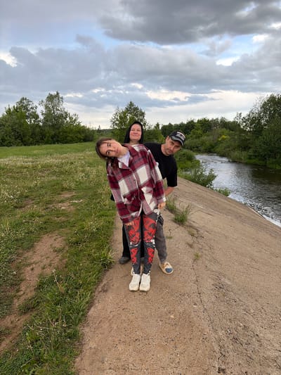 The image shows three people outdoors near a body of water, possibly a river. A man in a black t-shirt and a baseball cap is standing on a concrete structure, slightly behind a woman wearing a black hooded garment. In front of them is a younger woman wearing a red and white plaid jacket and pants with a graphic design. They appear to be posing for a photo. The location is identified as Nizhnii Vishur, Russia. The background includes trees, grass, and a cloudy sky.
