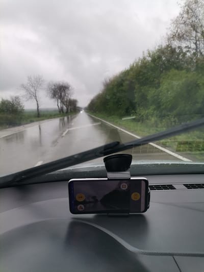 The image captures a first-person perspective from inside a vehicle, looking out through the windshield onto a wet, rural road. The scene is set on an overcast day, with a grey, cloudy sky dominating the upper portion of the frame. The asphalt road, glistening with water from recent or ongoing rain, stretches into the distance, reflecting the muted light and surrounding trees. White road markings are visible, guiding the path.

On the left side of the road, a line of mostly bare trees, likely deciduous, stands against a flat, possibly agricultural field. On the right, a denser, greener embankment covered in lush bushes and trees suggests a wooded area or hillside. A single windshield wiper blade is prominently positioned across the middle of the view, indicating wet conditions. Far down the road, the faint headlights of an approaching or distant vehicle can be seen.

In the foreground, the car's dashboard is visible, textured and dark. A smartphone is mounted centrally on the dashboard in a black holder. The phone's screen displays a user interface with several icons and text: a prominent yellow circle with the number "219", a yellow warning triangle with an exclamation mark, and what appears to be navigation or mapping controls, including an open book icon, a scan/focus icon, and a location pin icon.

The overall atmosphere is cool and damp, characteristic of a rainy day in a rural setting, possibly in Slaveevo, Bulgaria, given the provided context. No people are visible in the image.