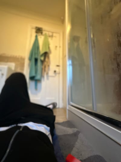 The image displays an interior domestic scene, likely a bathroom or an adjacent room, located in a residence in Dorchester, United Kingdom. The perspective is low, suggesting the camera is positioned on the floor. In the immediate foreground, the lower body and legs of an individual are visible, lying horizontally on a grey, textured surface, possibly a rug. The person wears dark trousers with a visible vertical zipper or seam and a white undergarment. One foot, wearing a grey shoe or slipper, is directed towards the background.
To the right, the lower section of a shower enclosure is present, featuring a silver-toned frame and glass panels that exhibit condensation or residue. A small red object rests on the grey surface near the shower's base. The midground shows a light-colored wall, revealing some damage or texture on its lower portion. A white door, equipped with a dark handle, stands in the background. A multi-hook hanger is mounted on the door's top edge, from which two bathrobes or large towels hang: one teal-blue and one light-green. A third lighter-colored garment is partially visible behind the green one. The lighting indicates an indoor setting. The scene is static, with no discernible actions or interactions beyond the individual lying on the floor and the arrangement of objects.