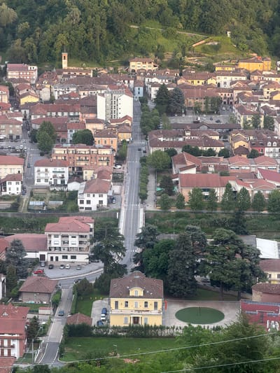 The image shows an aerial view of a section of Santo Stefano Belbo, Italy. At the upper left is a densely wooded hillside. Adjacent to the woods is a row of closely packed buildings with red tile roofs. A main road runs horizontally through the center of the image, with buildings and trees lining both sides. To the right, a circular area with a green surface is surrounded by trees and buildings. Cars are seen parked and driving on the roads. The overall environment is a mix of residential and commercial buildings interspersed with green spaces.