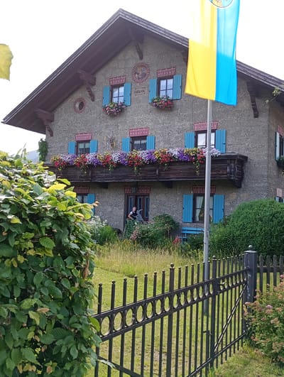 The image shows a multi-story building with a gabled roof, likely a house, in Brannenburg, Germany. The building has a stone facade, brown wooden window frames, and vibrant blue shutters. Numerous flower boxes overflow with colorful flowers on the balconies and window ledges, adding a cheerful aesthetic. A wooden balcony juts out from the second story, adorned with a dense arrangement of flowers. A flag, colored yellow and blue, flies from a pole in front of the building. In the foreground, a dense green bush partially obscures the view of the house. A black metal fence runs along the bottom of the image, separating the yard from the viewer. A young girl is visible standing near the entrance of the house, holding a toy or tool. The surrounding area appears to be a garden or yard with tall grass and other greenery.