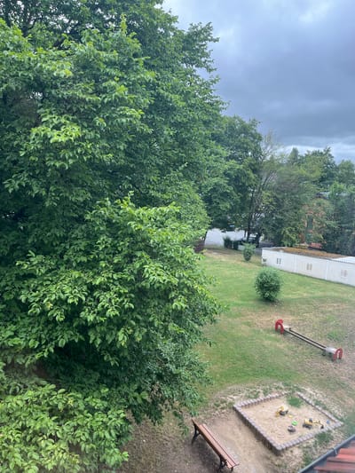 This is an aerial outdoor shot of a small, grassy playground and sandbox area adjacent to a building with a sloped, tiled roof. The playground features a sandbox with several toy construction vehicles, a wooden bench, and a see-saw with red arches. Beyond the playground, there's a grassy lawn with a few small trees and bushes. In the background, a white building with several small windows is visible, along with more trees and buildings, and a road with a parked car. The sky is overcast and cloudy, suggesting a possibly rainy day. The overall scene appears to be located in a residential area, likely a communal space within an apartment complex or a small public park. The image is taken from an elevated perspective, looking down at the playground and surrounding area.