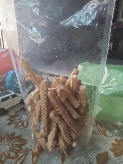 A clear plastic bag filled with brown, textured, stick-like snacks is sitting on a table with a floral patterned tablecloth. A toy car and a blue toy are visible beside the bag. A green plastic bag is behind the bag. The location is Kruengmanyang, Indonesia.