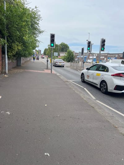 A street scene in Stoke-on-Trent, United Kingdom, features a white car stopped at a traffic light. The image is oriented vertically, with the sky at the top and the ground at the bottom. Several traffic lights are visible, with some showing a green signal. Buildings line the street, including one with "EDGEWOOD ACADEMY" written on it. People are seen walking on the sidewalk in the distance, some wearing high-visibility vests. The weather appears to be overcast, and the time of day seems to be daytime. The car has yellow stickers on its side, one of which appears to be advertising a taxi service. The road is asphalt with white lane markings. Green trees and foliage are visible on the left side of the image.